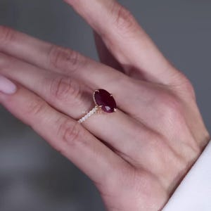 Puede incluir: Un anillo con una gran piedra preciosa de color rojo intenso de corte ovalado montada en una banda de oro. La banda está adornada con una hilera de pequeñas piedras transparentes engastadas en pavé a lo largo del aro. La palabra MARLETTE está grabada.