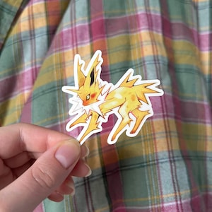 May include: A yellow and orange sticker depicting a Jolteon character. The sticker has a white border and is held against a plaid background with pink, green, and yellow tones.