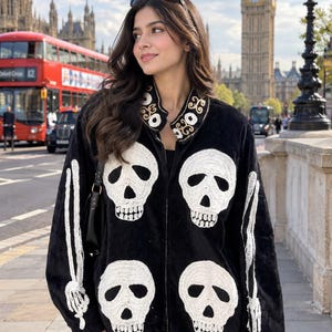 May include: A black zip-up jacket featuring large white embroidered skull motifs on the front and sleeve details resembling arm bones. The collar is accented with a decorative white and gold embroidered pattern, and the jacket has a soft texture.