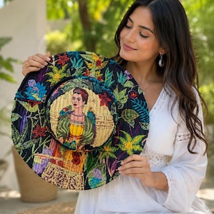 May include: A wide-brimmed quilted hat featuring a black base with vibrant, multi-colored floral and botanical embroidery. The center of the hat displays a detailed portrait of a woman with dark hair, wearing a yellow top and green shawl.