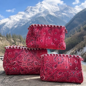 May include: Three quilted pouches in varying sizes, each featuring a vibrant red fabric with a dark paisley pattern. The pouches have a rectangular shape, a top zipper closure, and a decorative red and white striped trim along the upper edge.