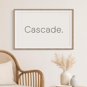 Op de afbeelding: Een ingelijste print met het woord "Cascade" in een minimalistisch lettertype hangt boven een rotan stoel met een crèmekleurig kussen. Een kleine houten bijzettafel bevat een vaas, een kleine keramische fles en een bord.