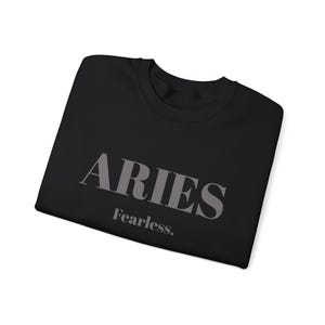 Puede incluir: Sudadera negra de cuello redondo con la palabra "ARIES" en gris y la palabra "Fearless." debajo. La sudadera está doblada. El diseño está centrado en la parte delantera de la prenda.