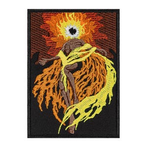 May include: Embroidered patch featuring a stylized figure with fiery wings and a halo-like element. The design uses shades of brown, orange, and yellow against a black background. The patch is rectangular and likely for clothing or accessories.