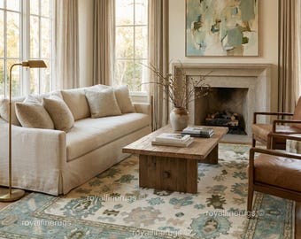 May include: A living room scene featuring a beige sofa with throw pillows, a wooden coffee table, and two leather armchairs. A large rug with a floral pattern in shades of blue, green, and beige covers the floor. An abstract painting hangs above a fireplace.