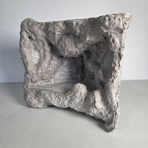 Może przedstawiać: Teksturowana, szara, abstrakcyjna rzeźba z wnętrzem przypominającym jaskinię. Dzieło ma szorstką, nierówną powierzchnię i prostokątny kształt. Rzeźba wykonana jest z jasnoszarego materiału i stoi na białej powierzchni.