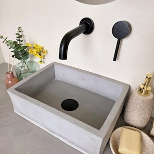 Könnte beinhalten: Ein rechteckiges Waschbecken aus Beton mit schwarzer Armatur und Griff. Ein Stück Seife liegt auf einer kleinen, gesprenkelten Schale. Ein dekorativer Seifenspender und eine Blumenvase sind ebenfalls zu sehen.