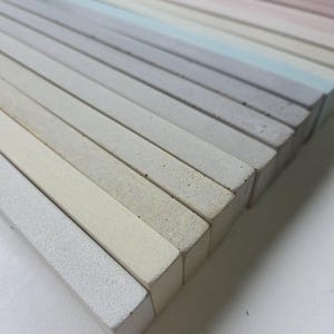 Può includere: Una collezione di bastoncini rettangolari in cemento color pastello disposti in file parallele. I colori variano dal rosa chiaro a varie sfumature di grigio e blu, creando un effetto sfumato. Le estremità dei bastoncini sono visibili.