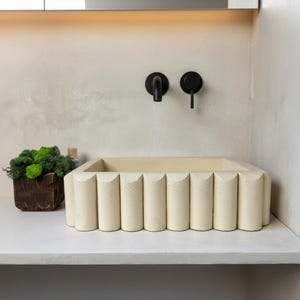 Peut inclure: Un lavabo rectangulaire crème avec un design cannelé. Le lavabo est posé sur un comptoir blanc, avec des robinets noirs fixés au mur au-dessus. Un pot en bois avec de la mousse verte est à gauche.