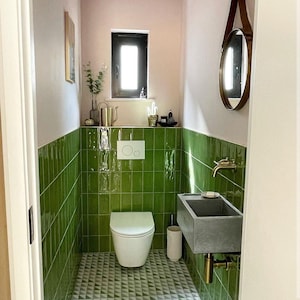Pode incluir: Uma casa de banho moderna com paredes de azulejos verdes e um chão com padrão geométrico. A casa de banho tem uma sanita branca, uma pia de cimento com acessórios de latão e um espelho redondo. Uma pequena janela fornece luz natural.