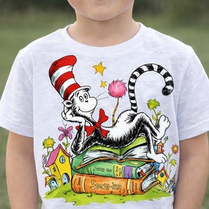 May include: A white t-shirt featuring a colorful illustration of the Cat in the Hat character from Dr. Seuss. The Cat is lounging on a stack of books, wearing his signature red and white striped hat and bow tie. The shirt is worn by a young child.