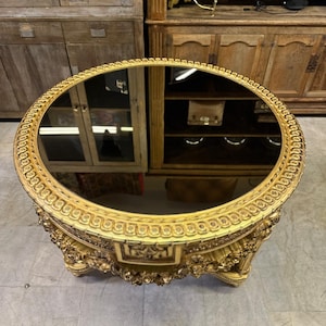 May include: Ornate gold-colored oval table with a mirrored top. The table features detailed carvings and a decorative edge. The table has three ornate legs and is likely an antique or vintage piece.