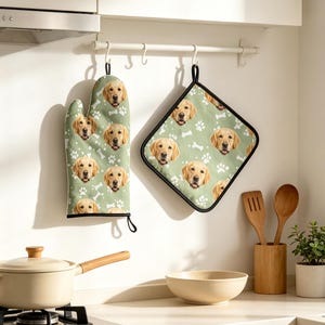 May include: A sage green oven mitt and a square pot holder featuring a repeating pattern of golden retriever dog faces and white paw prints. Both kitchen accessories have black trim and are hanging from a white wall-mounted rack.
