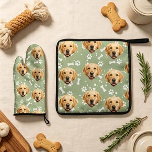 May include: A kitchen oven mitt and a square pot holder featuring a repeating pattern of golden retriever dog faces, white paw prints, and white bone shapes on a sage green background, both finished with black fabric trim and a hanging loop.