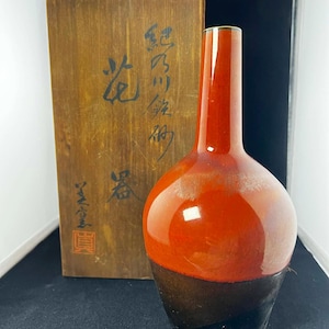 Peut inclure: Un vase en céramique bicolore avec une finition brillante. La partie supérieure est orange vif, passant à une base marron foncé. Une boîte en bois avec des caractères japonais est derrière le vase.