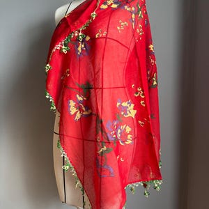Può includere: Un foulard rosso vivace caratterizzato da un motivo floreale con petali gialli e viola e foglie verdi. I bordi del foulard sono rifiniti con un delicato e complesso bordo in pizzo lavorato all'uncinetto nei colori verde e bianco.