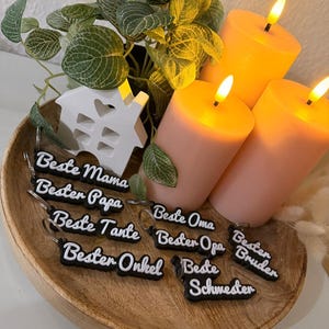 May include: A wooden tray displays black keyrings with white text, such as "Beste Mama" and "Bester Papa." Three lit, peach-coloured candles and a small white house ornament are also visible, alongside some greenery.