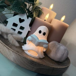 Könnte beinhalten: Eine Halloween-Dekoration mit einer weißen Geisterfigur, die ein Buch liest, einem kleinen weißen Haus mit Herz-Ausschnitt und einem glitzernden Kürbis. Die Anordnung umfasst drei brennende Kerzen auf einem Holztablett.