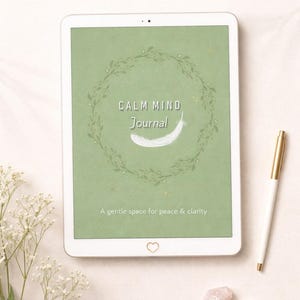 May include: A digital tablet displaying a journal cover with the text "Calm Mind Journal" and a feather illustration. The cover is surrounded by a green wreath. A white and gold pen rests beside the tablet, with small crystals and flowers in the foreground. The text "A gentle space for peace & clarity" is also visible.