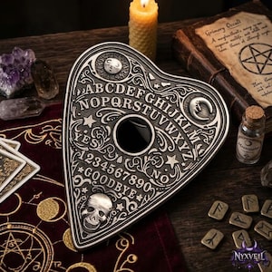 Könnte beinhalten: Eine herzförmige, silberfarbene Planchette mit geprägtem Alphabet, den Zahlen null bis neun, den Wörtern YES, NO und GOODBYE, einer zentralen runden Öffnung, einem Totenkopfmotiv sowie Himmelsymbolen wie Sonne und zunehmendem Mond.