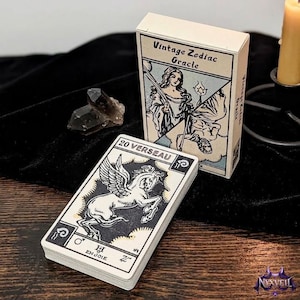 Könnte beinhalten: Ein Kartendeck Vintage Zodiac Oracle mit einer Schachtel, die eine Figur mit Stab zeigt. Eine Karte zeigt ein geflügeltes Pferd mit dem Text 20 VERSEAU und EN JOIE. Ein dunkler Kristall und eine gelbe Kerze in einem schwarzen Halter liegen daneben.