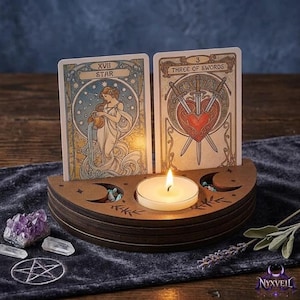 Puede incluir: Un portavelas de madera semicircular con dos ranuras para cartas de tarot, que presenta una vela de té encendida en el centro. Las cartas mostradas son La Estrella y el Tres de Espadas. El soporte tiene fases lunares y motivos de hojas grabados.