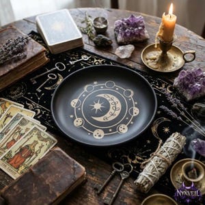 May include: A round black ceramic plate featuring a gold-colored print of a central sun and crescent moon surrounded by a lunar phase cycle. The plate is positioned on a dark cloth decorated with gold celestial patterns, tarot cards, and crystals.