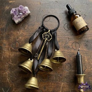 May include: A black metal ring adorned with four brass bells and a decorative brass skeleton key, all suspended by black satin ribbons. Nearby are a purple amethyst crystal cluster, a brown glass bottle labeled AURA OIL, and a black candle.