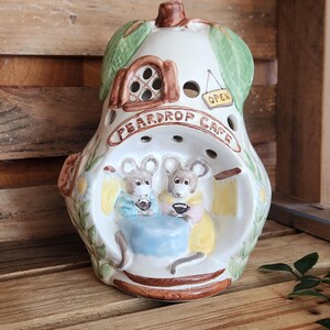 May include: A pear-shaped ceramic candle holder with the words "PEARDROP CAFE" and two cartoon mice. The cafe has an open sign and a green leaf design. The candle holder is on a wooden surface.