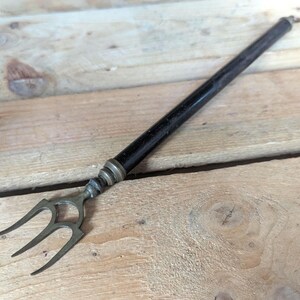 May include: An antique serving fork with a dark wooden handle and a three-pronged metal head. The handle is black with silver-colored metal accents near the head. The fork is set against a weathered wooden surface.