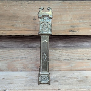May include: A vintage brass-colored miniature grandfather clock. The clock features a detailed face, a long rectangular body with a diamond-shaped accent, and decorative top and bottom elements. The clock is displayed against a wooden background.