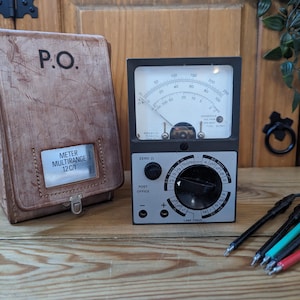 May include: A vintage multimeter with a black and silver face, a needle gauge, and a dial. A brown leather case with "P.O." and "METER MULTIRANGE 12 C/1" is on the left. Test leads with red, green, and black tips are in the foreground.