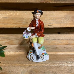 May include: A porcelain figurine of a person wearing a dark red jacket, yellow breeches, white stockings, and a black hat. The figure sits on a tree stump base with painted floral details, wearing black shoes and a white cravat at the neck.