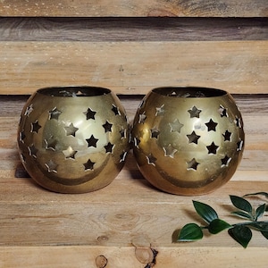 May include: Two spherical, gold-colored candle holders with star-shaped cutouts. The candle holders are made of metal and sit on a wooden surface. Green leaves are in the bottom right corner.