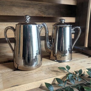 May include: Two shiny silver coffee pots with lids and curved handles sit on a wooden surface. The pots are reflective, showing the surrounding environment. The pots are of different sizes, suggesting a set or collection.