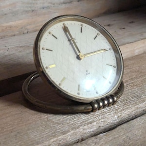 May include: A round, metallic travel clock with a cream-colored face featuring a subtle diamond pattern. The clock has gold-tone hands and hour markers, with the text 8 DAY printed on the dial. It is supported by a circular, hinged metal base.