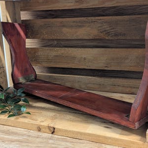 May include: A dark red wooden shelf with a curved design. The shelf is supported by two uprights and has a flat surface for displaying items. The shelf is placed inside a wooden crate.