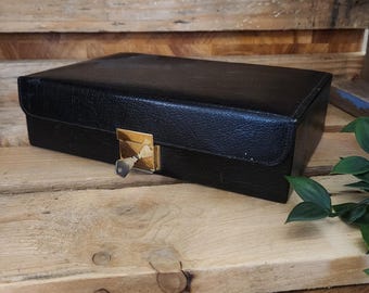 May include: A black rectangular box with a gold-colored clasp and key. The box is made of a textured material and sits on a wooden surface. The box is closed and appears to be vintage.