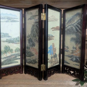 May include: A four-panel folding screen featuring a landscape painting. The dark wood frame has decorative cutouts at the base and brass hinges. The artwork depicts mountains, trees, and water in muted colours.