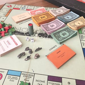 May include: A Monopoly board game set with various components. Includes stacks of colorful play money, dice, game tokens, Chance and Community Chest cards, and wooden houses and hotels. The board game is on a light green surface.