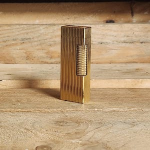 May include: A rectangular gold-tone metal lighter featuring a vertical ribbed texture across its body and a circular ignition roller on the side. The lighter has a clean, metallic finish with a consistent pattern of fine lines running from top to bottom.