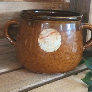 May include: A brown ceramic pot with two handles and a rounded body. The pot has a speckled finish and a label on the front. The label reads "Traditional French Cookware".