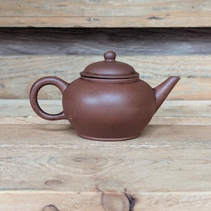 May include: A small, brown ceramic teapot with a rounded body, a curved handle, and a spout. The lid has a small knob on top. The teapot is sitting on a wooden surface.