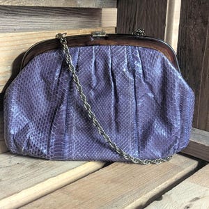 May include: A vintage, purple clutch bag with a snakeskin pattern. The bag has a wooden frame and a silver chain strap. The clasp is silver. The bag is on a wooden surface.