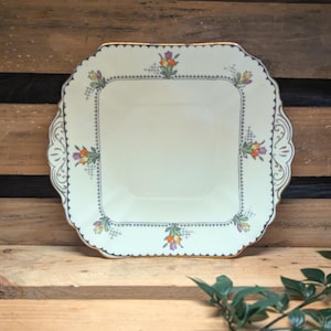 May include: Square, off-white vintage serving plate with a scalloped edge and gold trim. The plate features floral designs in orange, purple, and yellow. The plate is set on a wooden surface.