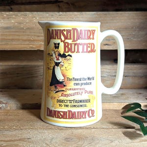 May include: A cream-colored ceramic pitcher featuring a vintage-style graphic of a person carrying a bucket. The text reads: DANISH DAIRY BUTTER, The Finest the World can produce, GUARANTEED ABSOLUTELY PURE, DIRECT FROM THE FARMHOUSE TO THE CONSUMER, DANISH DAIRY CO.