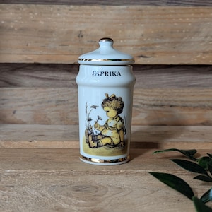May include: A white ceramic spice jar with a lid, featuring a gold rim and the word "PAPRIKA" in black. The jar is decorated with a vintage illustration of a child. The jar is approximately 4 inches tall.