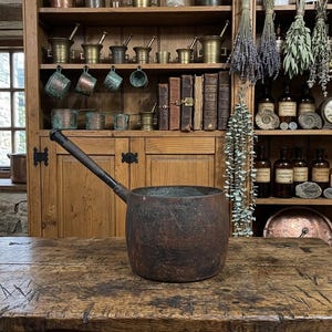 May include: A rustic, cylindrical metal pot with a dark, weathered, and oxidised brown finish. The pot features a single, long, straight handle extending from the side. The surface shows signs of age, including texture and discolouration.