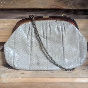 May include: A vintage light grey clutch bag with a snakeskin pattern. The bag has a brown tortoiseshell frame and a chain strap. The bag is made of a textured material and has a clasp closure.