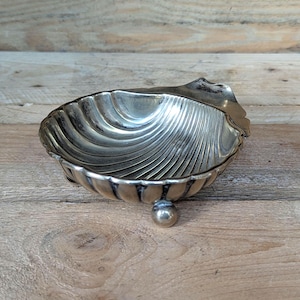 May include: A silver-colored, shell-shaped dish with a ribbed interior and a small, round foot. The dish has a scalloped edge and sits on a wooden surface. The dish is likely made of metal and is designed for decorative purposes.